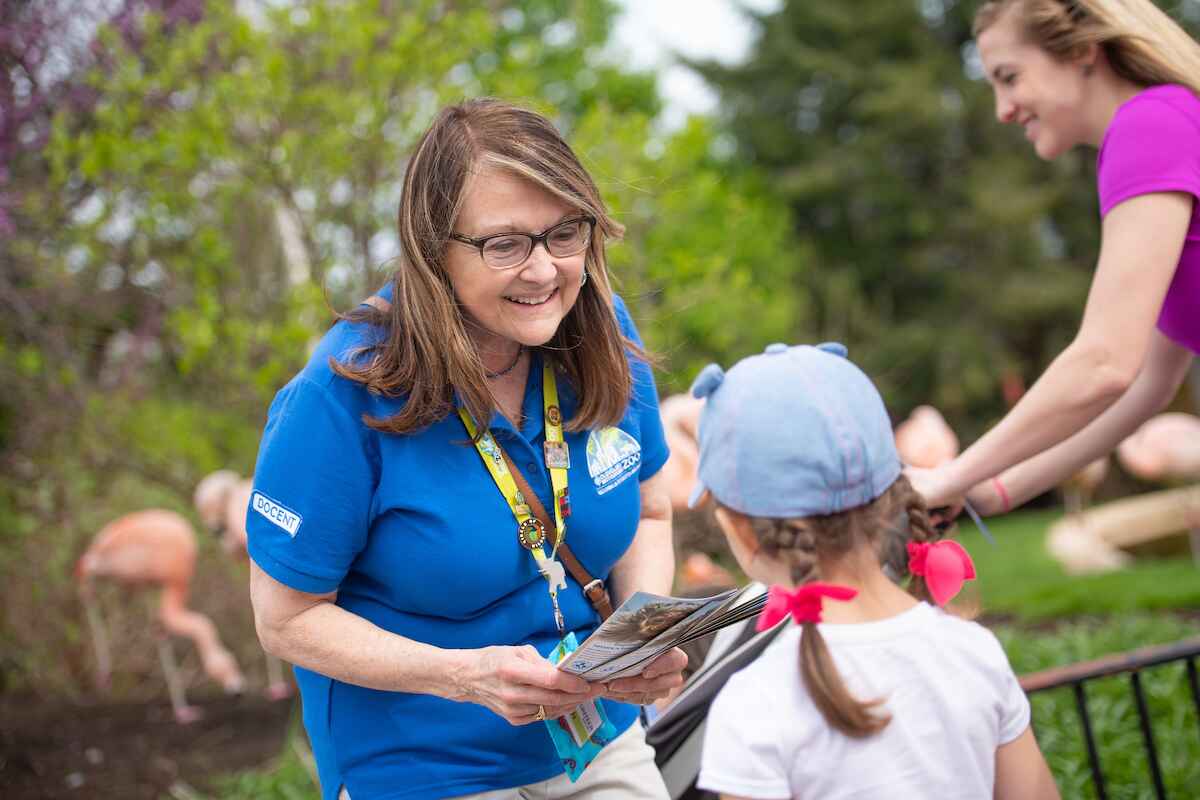 A smiling zoo ambassador in a blue shirt interacts with a young girl, with flamingos visible in the background.