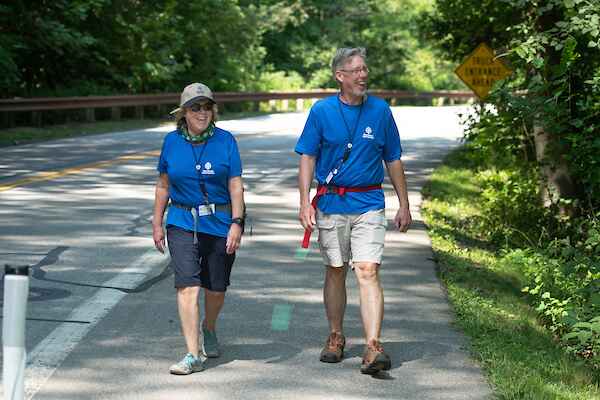 Two people wearing blue shirts and outdoor gear walk along a shaded road on a sunny day.