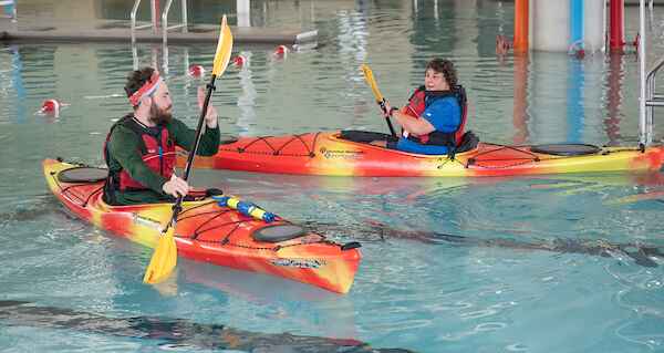 Two people in kayaks paddle in an indoor pool, wearing life jackets and holding paddles.