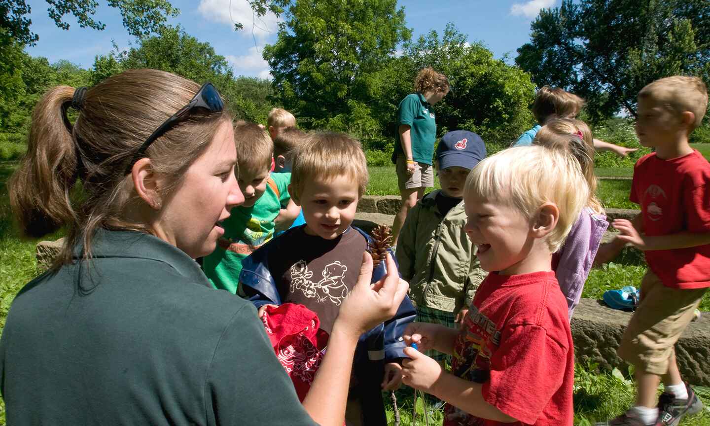 A group of young children outdoors, observing a pine cone held by a guide.