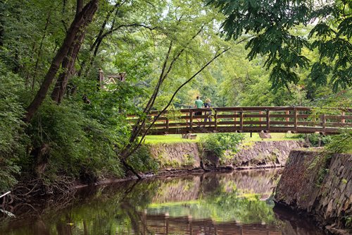 there is a man walking across a bridge over a river, an impressionist painting by Jeff A. Menges, featured on cg society, barbizon school, parks and public space, old stone bridge over the creek, woodland creek