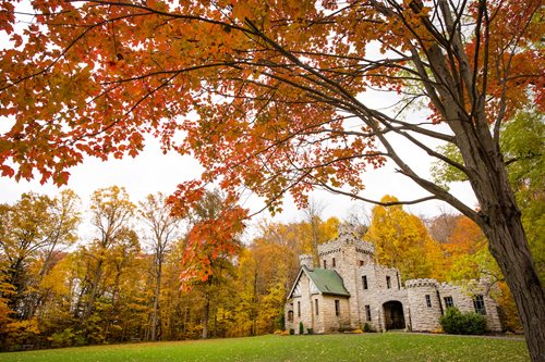 A stone building church school in an open field in the fall time with orange red green yellow leaves surrounding it, a castle like building