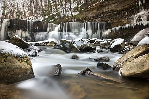 A serene waterfall cascades over rocks in a snowy woodland setting, surrounded by lush greenery and tranquil nature.