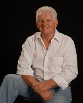 Man with white hair in a white shirt, seated with hands resting on his knee, smiling in front of a black background.