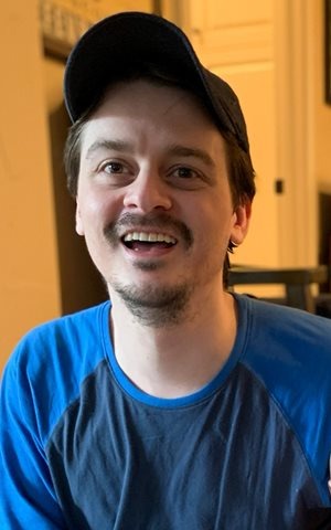 Man with a baseball cap and a mustache, smiling indoors, wearing a blue and black shirt.