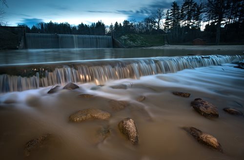 Water flows gently over rocks in a river, bordered by trees, illustrating a peaceful and scenic outdoor environment.