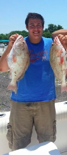 Young man holding two large fish, smiling proudly in a blue shirt on a sunny day.