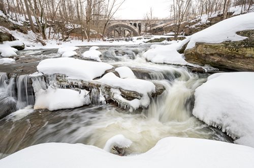 A picturesque river flows gently through a snowy forest, with rocks and trees framing the peaceful winter scene.