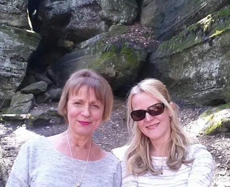 Two women smiling outdoors in a rocky, shaded area, one wearing sunglasses and the other with short hair.