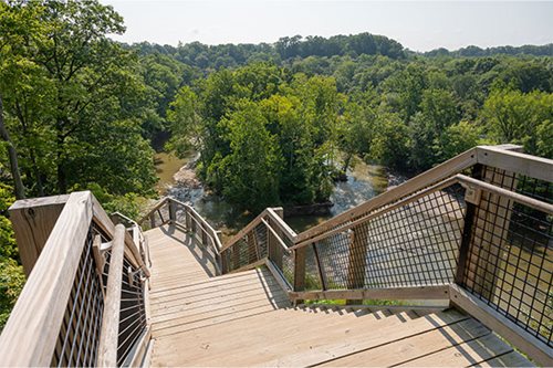 A wooden bridge with a railing spans across a serene river, surrounded by lush greenery and calm waters.