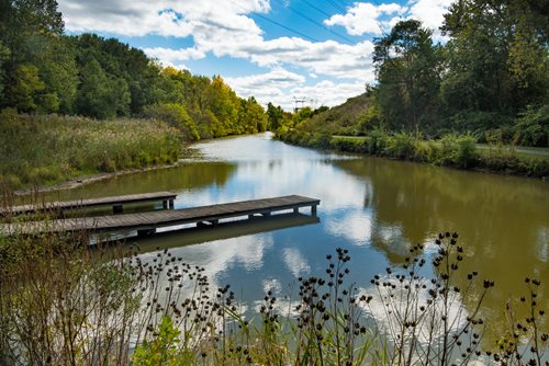 A small dock extends over the calm waters of a river, surrounded by lush greenery and a clear blue sky.