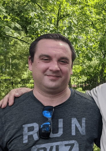 Man smiling outdoors, wearing a dark t-shirt with sunglasses tucked into his shirt, with greenery in the background.