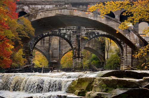 a stone bridge above a rushing waterfall with bright yellow leaves on the rightside and red leaves alongside the left 