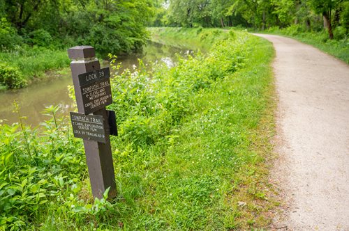 Scenic view of the Ohio & Erie Canal Towpath Trail at Brecksville, featuring a lush green landscape, a tranquil canal, and a clear walking path next to a Lock 39 signpost, ideal for hiking and nature exploration.