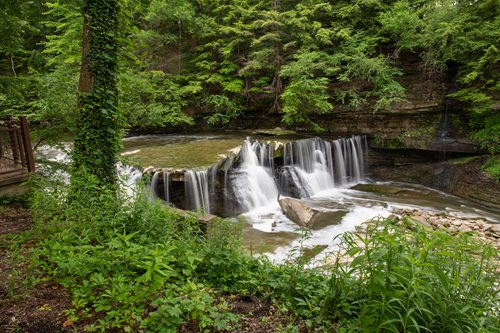 A serene waterfall cascades through the woods, accompanied by a rustic wooden walkway for visitors to enjoy the scenery.