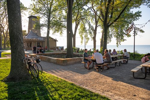 people sitting at picnic tables in a park near the water, a stock photo by Aaron Bohrod, instagram contest winner, regionalism, parks and public space, un restaurant avec une terrasse, parks and lakes