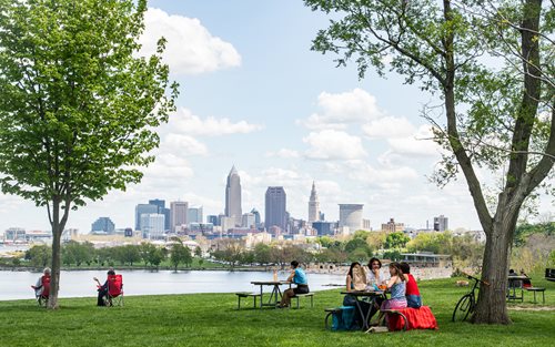 people sitting at picnic tables in a park with a view of the city, a stock photo by Joe Stefanelli, shutterstock contest winner, regionalism, parks and public space, parks and lakes, cleveland